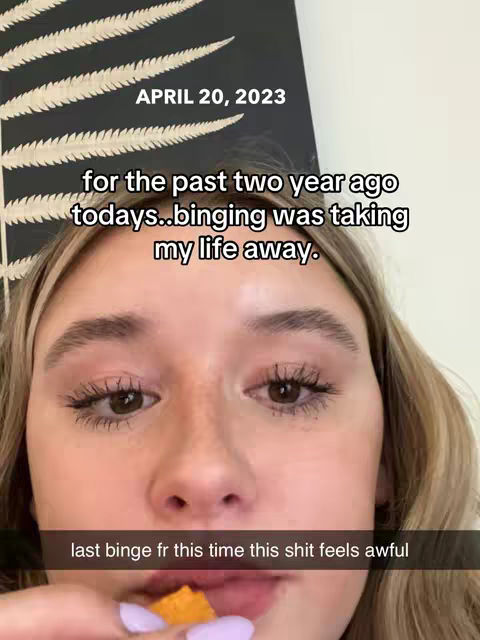The image shows a close-up of a young woman's face. She has fair skin, light-colored hair and is looking directly at the camera. A snack is in her hand. The background is a floral pattern. The text overlays include "APRIL 20, 2023", "for the past two year ago todays..binging was taking my life away.", and "last binge fr this time this shit feels awful."