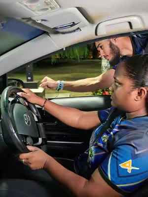 A scene inside a car shows a woman in the driver's seat with a man leaning in from the front passenger side, extending his arm as if pointing or gesturing toward something outside of the car. The background is blurred but suggests an outdoor setting.