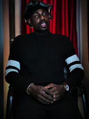A man with dark skin and a serious expression sits in a chair. He is wearing a black sweater with white stripes on the sleeves and a black hat. His hands are clasped in front of him. The background consists of red curtains.