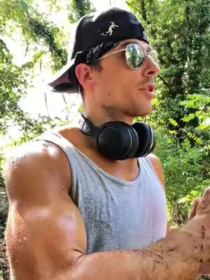 A medium shot features a tanned man with a muscular build against a blurry outdoor backdrop. He wears a gray tank top, a black cap with a design, and mirrored sunglasses, with a set of black headphones draped around his neck. He appears to be speaking, with his mouth slightly open. The lighting suggests a sunny day, and the background is filled with green foliage.