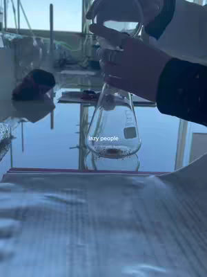The video shows a person wearing a lab coat, holding a flask with a clear liquid inside. The flask has a small amount of material at the bottom, and the text overlay 'lazy people' is visible on the flask. The background shows a window and a reflective surface, possibly a lab bench.