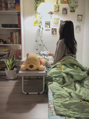 A young woman is seen from the back, sitting on a bed with a green blanket partially covering her. A small desk or table with a Winnie-the-Pooh plushie sits in front of her, and above her is a wall decorated with drawings and plants. The room is softly lit, giving a cozy and inviting feel.