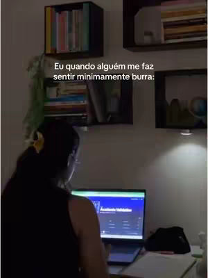 The image shows a person, likely a student, at a desk with a laptop. The person, seen from the back, is wearing glasses and has their hair up in a bun. The setting includes shelves filled with books and a wall with warm lighting. Text overlayed on the image reads, 'Eu quando alguém me faz sentir minimamente burra:'.