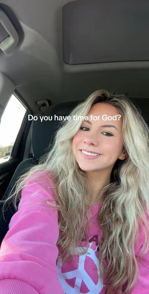 A young woman with long blonde hair is seated in a car. She's wearing a pink sweatshirt with a peace sign, and the text overlay reads, "Do you have time for God?"