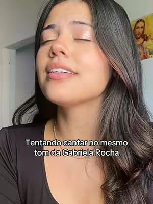 A young woman with long dark hair is shown with her eyes closed and mouth slightly open, perhaps singing or feeling emotion. Text overlay at the bottom of the screen says 'Tentando cantar no mesmo tom da Gabriela Rocha'. A religious image is faintly visible in the background.