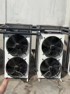 The image shows two large industrial-looking air conditioning units with four fans each, arranged in pairs. The units are white with black fan blades. The background is a light wall and a gray ground. A finger is in the bottom left corner.