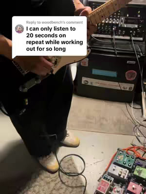 A partially visible shot shows a person standing on a gray floor, holding and playing a guitar. The person is wearing dark pants and light-colored shoes. In the foreground there is an array of guitar effects pedals connected. Behind them, there is an amplifier and other musical equipment. A text box which is a comment replies, is overlapping the guitar.