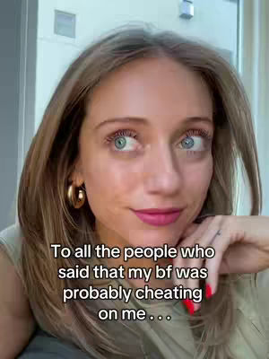 A selfie-style shot featuring a young woman with light brown hair and fair skin. She has light blue eyes, and a slight smile. Gold hoop earrings are visible. Pink lipstick and red nail polish are visible on her lips and fingers, and a small tattoo is on her left ring finger. The background seems to be an apartment or a house, as part of a white wall and window are present.