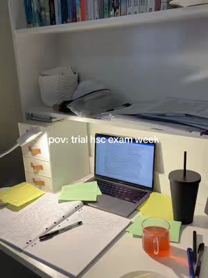 A cluttered desk with an open laptop, notebooks, and study materials. The background shows a bookshelf and a white shelf. A glass of red liquid is on the desk.