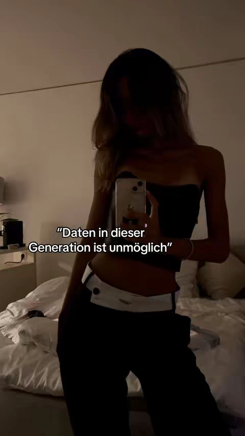 A person in a room is taking a selfie in front of a mirror with a phone. The person is wearing a black top and black pants, and the background is blurry.