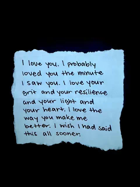 The image is a stark black background with a light blue torn piece of paper in the center. Handwritten in black ink on the paper is a message in a cursive-like font, expressing feelings of love and admiration. The paper's torn edges give it a tactile feel, and the overall composition is simple yet intimate.