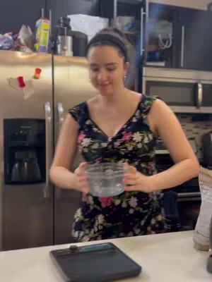 A woman in a floral dress is holding a clear bowl in a modern kitchen. A stainless steel refrigerator is beside her. A black digital device is visible on the counter in the foreground.