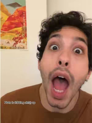A selfie-style shot featuring a young man with short, dark hair and a light complexion, his face close to the camera. His eyes are wide with surprise and his mouth is opened in a large, exaggerated expression. He wears a brown shirt. The background has elements out of focus, like the corner of a wall and a piece of wall art.