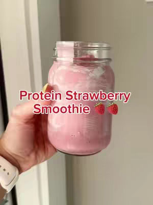A hand holds a mason jar filled with a pink smoothie. The jar has a silver lid and the text 'Protein Strawberry Smoothie' is overlaid in a pink font. Two cartoon strawberries are also added. The image is set against a neutral background.