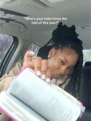 A selfie-style shot taken inside a car. The main subject is a Black woman with long, dark braided hair, wearing a brown top, and gold earrings. She's holding an open book (Bible), with one hand pointed towards it with white nail polish. The background is out of focus, showing the car's interior and rain on the window.