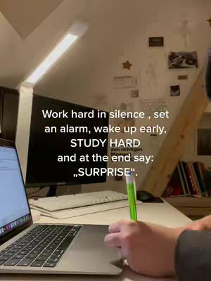A person is working at a desk. The foreground features a laptop and a hand holding a green pen, with a desk and light setup. The wall in the background has notes and drawings pinned to it.