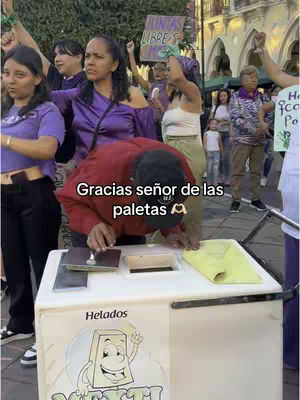 A group of people are gathered outdoors, with a person in a red jacket attending to an ice cream cart. The image is filled with people of various ages and genders, and the person in the red jacket is the focus, kneeling to serve from a white cart. The background includes other individuals and signs, possibly at a gathering or march. The image has a documentary style.