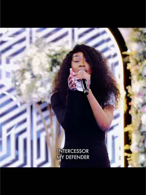 A woman with long, dark curly hair stands in front of a blurred background, singing into a microphone. The text overlayed says "INTERCESSOR MY DEFENDER."