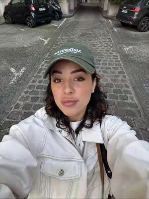 A selfie-style shot featuring a woman in a white jacket and a green cap, set against a background of a narrow, paved street with parked cars on either side. The woman is looking directly at the camera. The sky is overcast with shadows from the surrounding buildings.