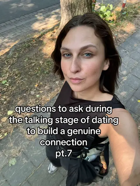 A woman with dark hair and blue eyes takes a selfie outdoors. The background includes trees, a sidewalk made of patterned pavers, and sunlight creating soft shadows. The woman is wearing a black top, and text overlay is in white, centered at the bottom.