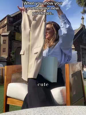 A woman is standing outdoors, partially obscured by a light-colored garment she holds up, with her arms raised. She's wearing a light blue button-up shirt and dark pants, posing near a wooden chair. The background includes a building with architectural details and a sunny, cloud-filled sky. The phrase 'When you show your partner your new purchases' is overlaid on the image, with the additional word 'cute' at the bottom.
