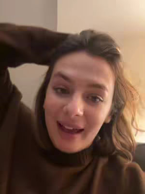 A woman is shown close-up, with her face filling most of the frame. She's looking up and to the left, seemingly mid-sentence, with a slight smile. Her hand is placed near her hair, and she appears to be indoors. Warm lighting and a neutral background suggest a casual setting. She is wearing a dark brown sweater.