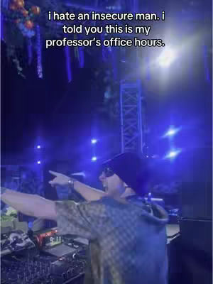 The image shows a person in a dark setting, likely a concert or club, with bright blue and white lights. The individual is wearing a dark beanie, sunglasses, and a patterned shirt, and is pointing with their right arm.
