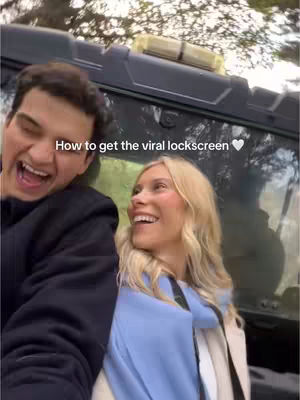 A selfie-style image shows a smiling couple against an outdoor background. The man on the left has dark hair and a black shirt and the woman has blonde hair blue jacket and beige coat. Text overlayed on the image reads 'How to get the viral lockscreen'.