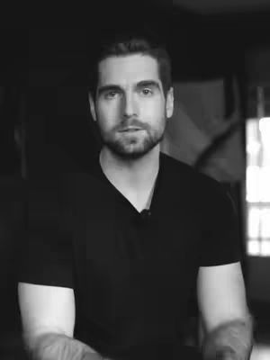 A grayscale portrait of a man in his late 20s or early 30s, with short, dark hair neatly combed. He has a trimmed beard and is wearing a black v-neck shirt. The background is dark, and the lighting is soft, focusing on his face and upper body. He is looking directly at the camera with a neutral expression.