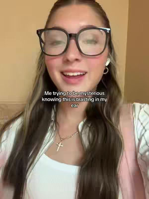 A young woman with long, multi-toned hair is wearing large, black-rimmed glasses and a white top, and a gold necklace with a cross. She's smiling with a slightly mischievous look, with text overlaid about being mysterious.