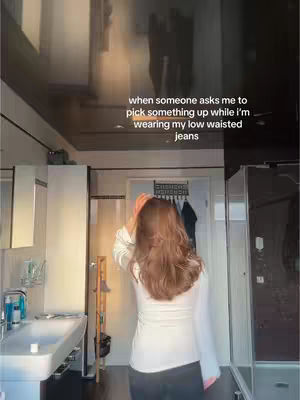 A bathroom scene with a woman standing facing away, wearing a white shirt and dark pants, reaching towards the door. Text is overlaid at the top.