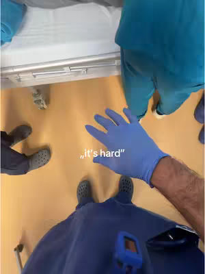 An overhead shot captures a medical setting. A person wearing medical scrubs and blue gloves extends their gloved hand towards the camera. The words "it's hard" are overlaid.