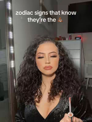 A woman with long, curly dark hair is centered in the frame. She has closed eyes and is holding a lipgloss applicator. The background is a softly lit room with a mirror and dresser visible.