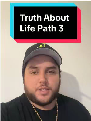 The image is a selfie-style shot featuring a man centered in the frame. He is wearing a black cap, black shirt and a gold chain. Behind the man is a light-colored neutral background. A rectangular text overlay is at the top of the frame, it displays white text on a black background with a teal and red border. The lighting appears to be soft and diffused, with no harsh shadows, suggesting an indoor setting.