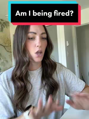 A woman with long brown hair poses for the camera. Above her head is a black rectangle with white text that reads "Am I being fired?". She is wearing a grey t-shirt.