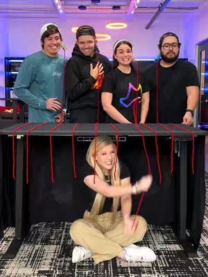 A group of five people standing and sitting around a black table with red strings attached to the table and connected to the hands of the individuals. A woman sits under the table, holding the strings. The scene is brightly lit with colorful lights and a plain background. The overall composition is dynamic, capturing a moment of interaction and potential storytelling.