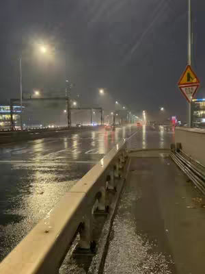 A nighttime shot of a wet street with illuminated streetlights lining the sides and reflections on the wet asphalt. A metal railing is in the foreground, with the road and distant lights creating a sense of depth.