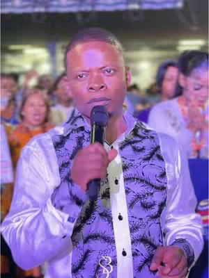 A man in a white and patterned shirt speaks into a microphone. The background is blurred but appears to be a gathering of people. The man is the focus, centered in the frame, and holding the microphone close to his mouth. The lighting is bright, and the color palette is dominated by purples and whites.