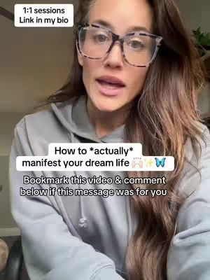 A woman wearing glasses and a gray hoodie is centered in the frame, speaking directly to the camera. Overlay text informs viewers to find 1:1 coaching links in the creator's bio, and also asks viewers to bookmark the video and comment below if the message resonates with them.