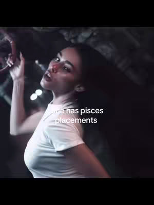 The cover image shows a woman looking up and to the left with a pose of surprise and her right arm raised. She is wearing a white t-shirt, and light shines from behind her, creating a dark, moody background. The words "She has pisces placements" are written in white font on the image.