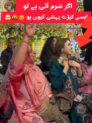 A group of people are gathered at a wedding or party. The image is dominated by a woman in a pink outfit, raising her arm and seeming engaged in conversation. A woman in green and gold outfit is smiling and looking to the right. There are flowers in the background, in addition to other people.