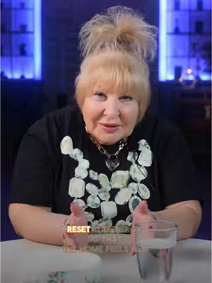 A medium shot. The main subject is a woman with blonde hair styled in a bun with bangs. She is wearing a black shirt with a white and green design. Her hands are up, to the side. The background is dark with some lights that look like blue bars and a table and a glass of water.
