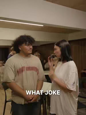 An interview setting, with two people standing and conversing. The background is a blurry room, suggesting a casual gathering or event. The bottom of the image has the hook text. The colors are natural, illuminated by overhead lights.
