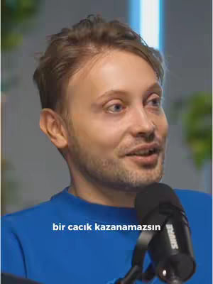 A man with light hair and a slight unshaven beard is speaking while holding a black microphone. Text at the bottom of the screen reads, "bir cacık kazanamazsın". The man wears a blue top.