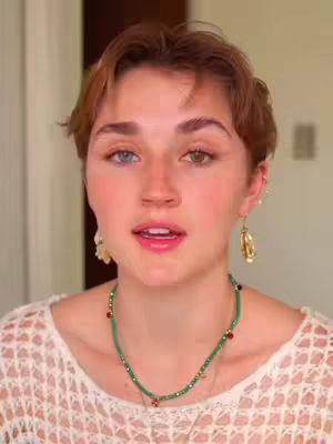 A woman with short brown hair and heterochromia (one blue and one brown eye) is centered in a medium shot. She wears a white, open-knit top and a green beaded necklace. Her mouth is slightly open as if speaking. Gold hoop earrings and a soft, neutral background complete the composition.