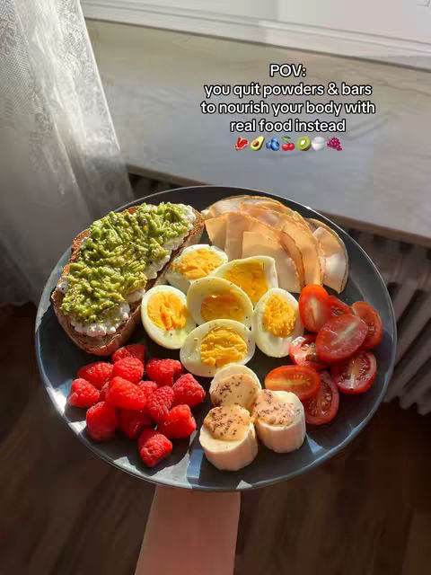 A plate of food with a variety of healthy items, arranged for a visually appealing presentation. The plate contains sliced tomatoes, halves of hard-boiled eggs, a pile of sliced deli meat, fresh raspberries, and slices of banana topped with nuts. The plate sits on a wooden surface, with a bright window in the background.