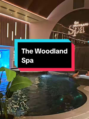 The cover image showcases an indoor spa scene, featuring a dark-colored pool in the foreground. A turquoise text box in the mid-ground with white text that reads 'The Woodland Spa'. Behind the pool is a darker semi-circular wall with a wooden panel backdrop, and a sign naming the spa. The ambient lighting of the scene gives a luxurious feel.