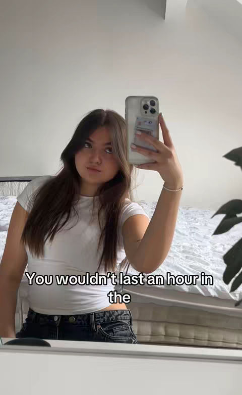 A young woman is taking a mirror selfie in what appears to be her bedroom. She's wearing a white t-shirt and dark jeans. Her long, dark hair is down, and she's holding a phone up with her right hand to take the photo showing the reflection of the woman and of a double bed. The background includes a mostly white wall and part of a bed with a white comforter.