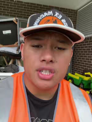 The cover image features a person wearing a hat with text on it, an orange vest, and ear piercings, with a building in the background. They are looking directly at the camera with an open mouth. There are some tools/lawn equipment on the side, next to the building.