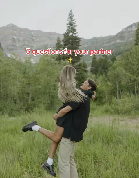 A couple is shown in a field with a mountain backdrop. The man is holding the woman up in his arms, and they are both smiling. The image is warm and inviting, with the green grass and trees surrounding them.